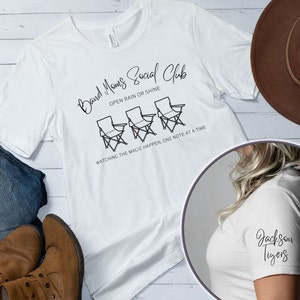 Peut inclure: T-shirt blanc avec du texte noir qui dit "Band Moms Social Club" avec trois chaises en dessous. Le texte continue "Open rain or shine" et "Watching the magic happen, one note at a time".