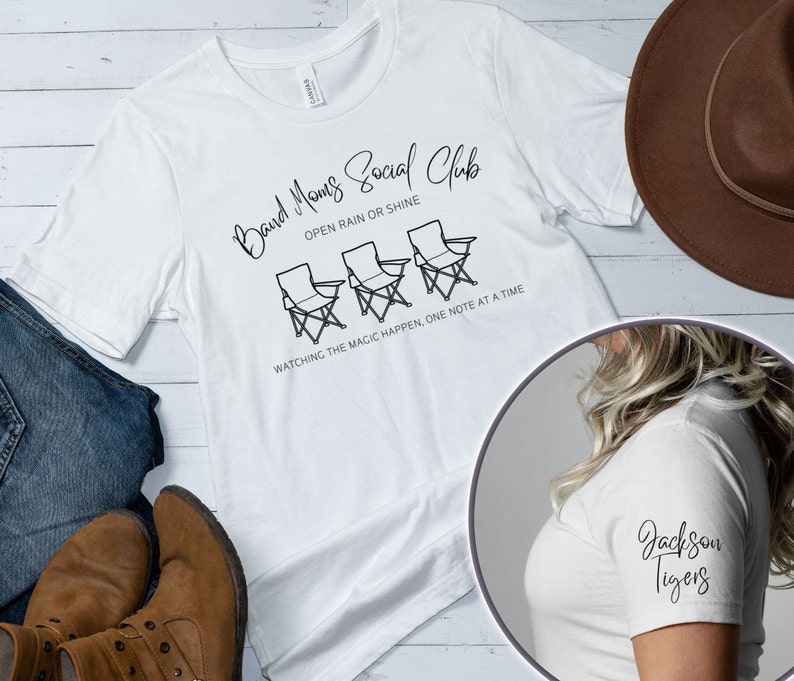 Peut inclure: T-shirt blanc avec du texte noir qui dit "Band Moms Social Club" avec trois chaises en dessous. Le texte continue "Open rain or shine" et "Watching the magic happen, one note at a time".