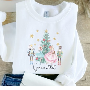 May include: White long-sleeved sweatshirt with a Christmas-themed design featuring a Nutcracker, a ballerina, a mouse king, and a Christmas tree. The text "Gracie 2023" is printed below the design.