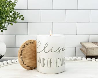 Puede incluir: Una vela blanca con una mecha encendida, con el nombre "Elise" y las palabras "Maid of Honor" en escritura gris. Una tapa de madera con la inscripción "Thank you for being my bestie" está al lado de la vela. La vela está sobre una superficie blanca.