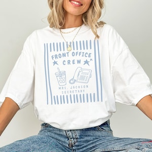 May include: A white t-shirt featuring a light blue graphic design with vertical stripes, the text "FRONT OFFICE CREW", "MRS. JACKSON SECRETARY", a drink cup, a landline telephone, and two stars. The shirt has short sleeves and a crew neckline.