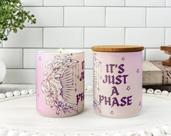 May include: Two cylindrical candles with a pink and purple gradient. One candle is lit, and the other has a wooden lid. The candles feature a crescent moon design and the phrase "It's Just A Phase" in purple.