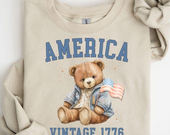 May include: Beige sweatshirt with the word "AMERICA" in blue block letters above a watercolor illustration of a teddy bear wearing a denim jacket and holding a small American flag. The text "VINTAGE 1776" is below the bear.