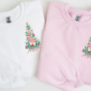 May include: Two Christmas-themed sweatshirts with a pink ballet shoe design on a green Christmas tree. The text "Ayana" is embroidered on each sweatshirt. One sweatshirt is white and the other is pink.