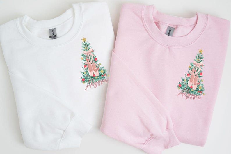 May include: Two Christmas-themed sweatshirts with a pink ballet shoe design on a green Christmas tree. The text "Ayana" is embroidered on each sweatshirt. One sweatshirt is white and the other is pink.