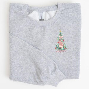 May include: A gray sweatshirt with a Christmas tree design featuring pink ballet shoes. The tree is decorated with red and green ornaments. The text "Merry & Bright" is embroidered below the tree.