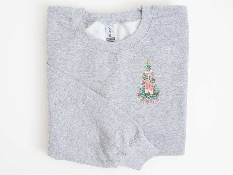 May include: A gray sweatshirt with a Christmas tree design featuring pink ballet shoes. The tree is decorated with red and green ornaments. The text "Merry & Bright" is embroidered below the tree.