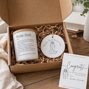 May include: A gift box containing a white candle with a wooden lid, a round white ceramic ornament, and a greeting card. The candle features a definition of White Coat. The ornament displays a white coat illustration, the name Kiley, and August 8, 2024. The card reads Congrats with a white coat graphic.