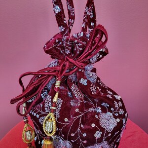 May include: A burgundy velvet drawstring bag with silver sequins and floral embroidery. The bag has a long tassel and a gold-colored metal embellishment.