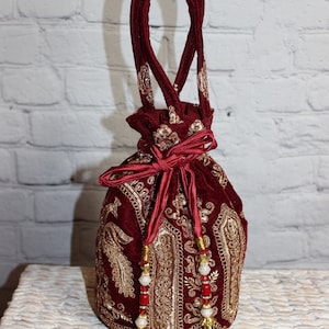 Może przedstawiać: Bordowa aksamitna torba ze sznurkiem i złotym haftem. Torba posiada pasujący bordowy uchwyt i detale z frędzlami. Złożony złoty haft pokrywa przód i boki torby, tworząc luksusowy wygląd.