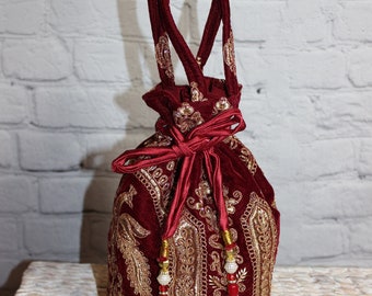 Könnte beinhalten: Ein bordeauxfarbener Samtbeutel mit goldener Stickerei. Die Tasche hat einen passenden bordeauxfarbenen Griff und Quasten. Die aufwendige goldene Stickerei bedeckt die Vorder- und Seiten des Beutels und sorgt für einen luxuriösen Look.