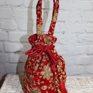 May include: Red velvet drawstring bag with gold floral embroidery and matching handles. The bag features a red satin bow closure and a tassel with gold beads. A decorative and elegant accessory.