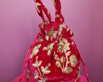 Könnte beinhalten: Ein roter Samtbeutel mit goldenen Blumenstickereien und Pailletten. Die Tasche hat einen geflochtenen Henkel und einen Kordelzugverschluss. Der Beutel steht auf einer rosa Oberfläche.