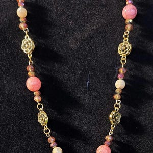 Può includere: Una collana dorata con perline rosa e crema, intervallate da perline iridescenti più piccole. La collana include anche dettagli decorativi dorati a forma di fiore, creando un design elegante e femminile.