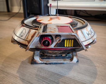 May include: A weathered, sci-fi prop with a circular design, featuring a white and gray body with rust-like accents. It has red and black details, two antennas, and the word "Kino" is visible. The prop sits on a matching base.