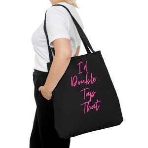 May include: Black tote bag with the phrase "I'd Double Tap That" in pink cursive lettering. The bag has long black straps and is carried over the shoulder. The bag is made of a sturdy material.