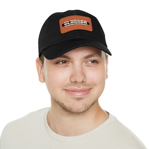May include: Black baseball cap with a brown leather patch. The patch features the word "CLOGGER" in white block letters. The cap is made of a durable fabric and has a curved brim.