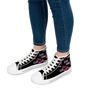 May include: Black high-top trainers with white soles and laces. The trainers feature the word "CLOG" in a repeating pink pattern. The shoes are worn with blue jeans.