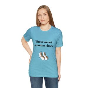 May include: A turquoise t-shirt featuring the phrase "These aren't wooden shoes" and an illustration of two pairs of white and brown shoes. The shirt is worn by a person.