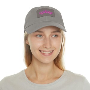 May include: A gray baseball cap with a curved brim. The cap features a rectangular patch with the word "BLOGGER" in pink, arched across the patch. The cap is made of a soft material and is suitable for everyday wear.