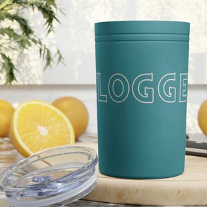 May include: Teal-coloured tumbler with the word "LOGGER" in white, and a clear lid. The tumbler is on a wooden chopping board with orange slices in the background. The tumbler is cylindrical and appears to be made of metal.
