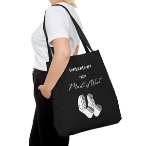 May include: Black tote bag with the text "Contents are NOT Made of Wood" in white script. The bag features a graphic of two pairs of white shoes with grey accents. The bag is carried over the shoulder.