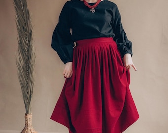 May include: A woman wearing a black long-sleeve blouse and a red skirt. The skirt is knee-length and has a full, pleated design. She is also wearing a red necklace and red ankle boots. The background is a neutral beige color.