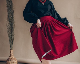 May include: A woman wearing a black blouse with a red beaded necklace and a red skirt. She is wearing burgundy ankle boots and is standing on a woven rug.