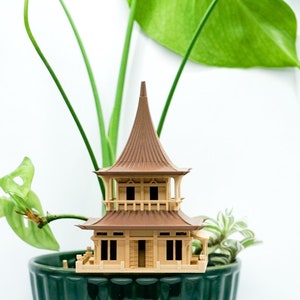 Può includere: Un piccolo modello di casa in legno marrone in stile pagoda si trova in un vaso di ceramica verde con una pianta verde frondosa che cresce.
