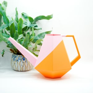 Könnte beinhalten: Eine geometrische Gießkanne mit facettiertem Design, bestehend aus einem hellrosa oberen Teil und einem leuchtend orangefarbenen unteren Teil, inklusive passendem orangefarbenem Griff und einem langen, schmalen hellrosa Ausguss.