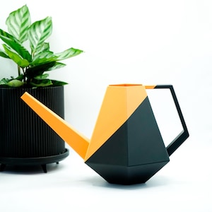Könnte beinhalten: Eine moderne, geometrische Gießkanne in Schwarz und Orange. Die Kanne hat einen langen, abgewinkelten Ausguss und einen schwarzen Griff. Im Hintergrund befindet sich eine Topfpflanze mit grünen Blättern.