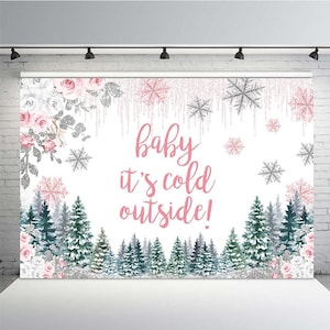 May include: A white backdrop with a pink and silver glitter design. The text "baby it's cold outside!" is written in pink cursive font. The background features a snowy forest with pink and white flowers.
