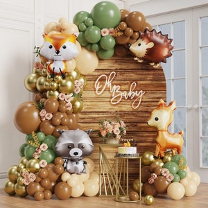 Op de afbeelding: Een babyborrel achtergrond met een bos thema met een houten bord waarop 'Oh Baby' staat, omringd door bruine, groene en gouden ballonnen. Er zijn ook ballonnen in de vorm van dieren, waaronder een vos, een egel, een wasbeer en een hert.