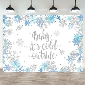 May include: A white backdrop with a winter theme, featuring blue and silver snowflakes. The phrase "Baby it's cold outside" is written in silver glitter script. Ideal for a baby shower or winter-themed event.
