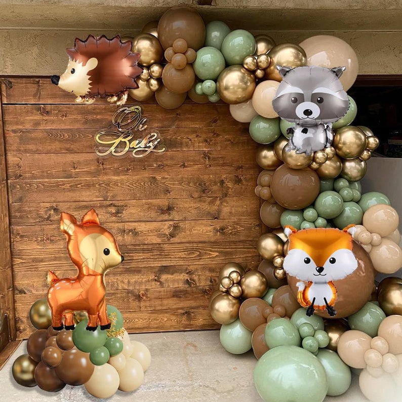 Op de afbeelding: Een baby shower achtergrond met een bos thema met een guirlande van bruine, groene en gouden ballonnen. De guirlande bevat dierballonnen, waaronder een vos, een wasbeer en een egel. De tekst "Oh Baby" wordt weergegeven op een houten achtergrond.