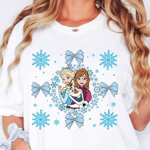 May include: White t-shirt featuring a colorful graphic of characters from the movie Frozen, including Elsa, Anna, and Olaf, surrounded by blue snowflakes and bows. The word "WHITE" is printed in the upper left corner.