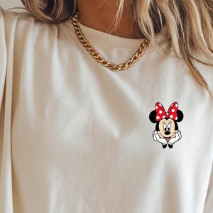 May include: Ivory-coloured t-shirt with a Minnie Mouse graphic. The graphic shows Minnie Mouse with a red polka-dot bow. The shirt has short sleeves and a crew neck. The word "IVORY" is printed at the top.