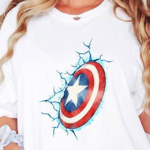 May include: White t-shirt featuring a Captain America shield design. The shield is red, white, and blue with a white star in the center. The design appears to be breaking through a cracked wall effect. The word "WHITE" is printed at the top.