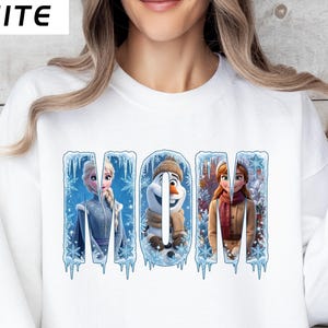 Puede incluir: Sudadera blanca con la palabra "MOM" en un dise&ntilde;o invernal, con personajes de una popular pel&iacute;cula de animaci&oacute;n. La palabra "WHITE" est&aacute; impresa en la esquina superior izquierda. El dise&ntilde;o incluye car&aacute;mbanos y copos de nieve.