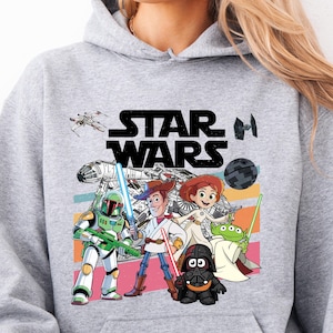 Puede incluir: Sudadera gris deportiva con un gr&aacute;fico colorido de Star Wars. El dise&ntilde;o presenta personajes como Darth Vader, Woody y Buzz Lightyear. El logotipo de Star Wars es visible. La sudadera tiene capucha con cord&oacute;n y bolsillo canguro.