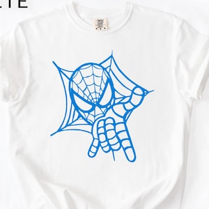 May include: White t-shirt with a blue graphic of a superhero's face and hand, surrounded by a spider web design. The word "WHITE" is printed in the upper left corner of the image.