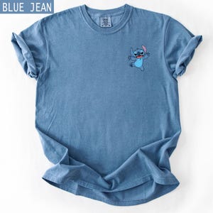 May include: Blue jean-colored t-shirt featuring a small embroidered image of the Disney character Stitch. The shirt has a crew neck and short sleeves. The text "BLUE JEAN" is in a blue rectangle in the upper left corner.