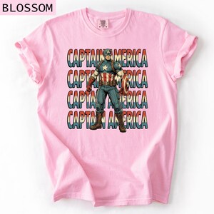 May include: A light pink t-shirt featuring a graphic of Captain America. The design includes the text "CAPTAIN AMERICA" repeated four times, with an image of Captain America in the center. The superhero is holding his shield.