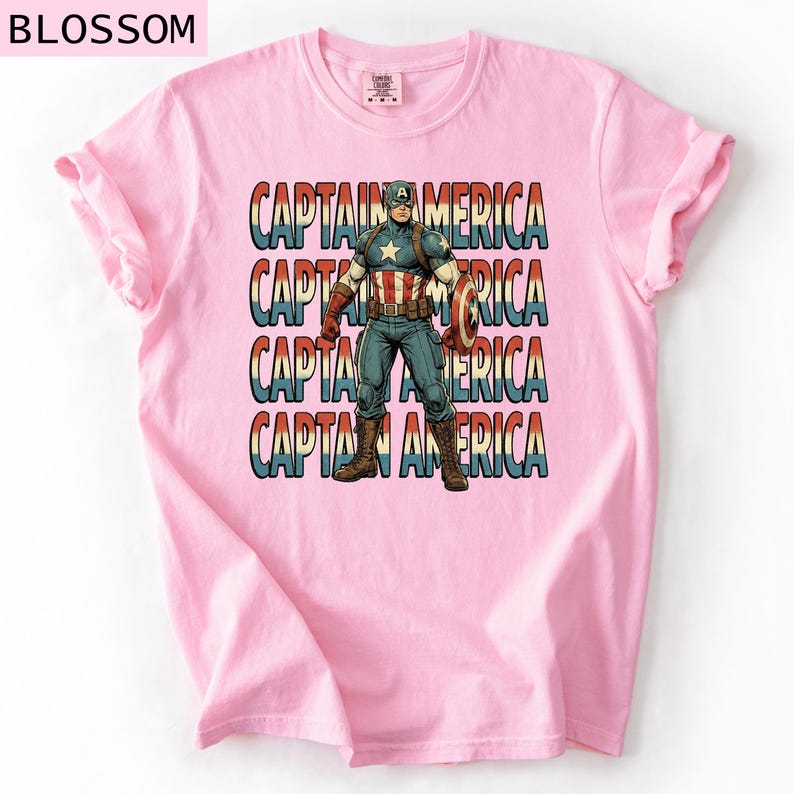 May include: A light pink t-shirt featuring a graphic of Captain America. The design includes the text "CAPTAIN AMERICA" repeated four times, with an image of Captain America in the center. The superhero is holding his shield.