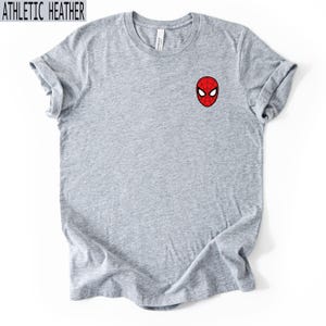 Puede incluir: Camiseta gris jaspeado con una peque&ntilde;a m&aacute;scara de Spider-Man roja y negra bordada en el pecho. La camiseta tiene mangas enrolladas y el texto "ATHLETIC HEATHER" en negro.