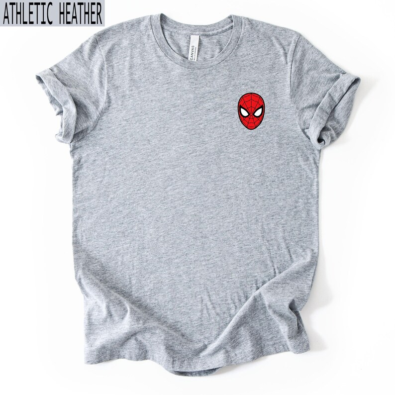 Puede incluir: Camiseta gris jaspeado con una peque&ntilde;a m&aacute;scara de Spider-Man roja y negra bordada en el pecho. La camiseta tiene mangas enrolladas y el texto "ATHLETIC HEATHER" en negro.
