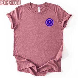 Pu&ograve; includere: T-shirt color malva melange con un disegno a scudo blu e beige sul petto. La maglietta &egrave; a girocollo e ha le maniche arrotolate. Il testo "HEATHER MAUVE" si trova nell'angolo in alto a sinistra.
