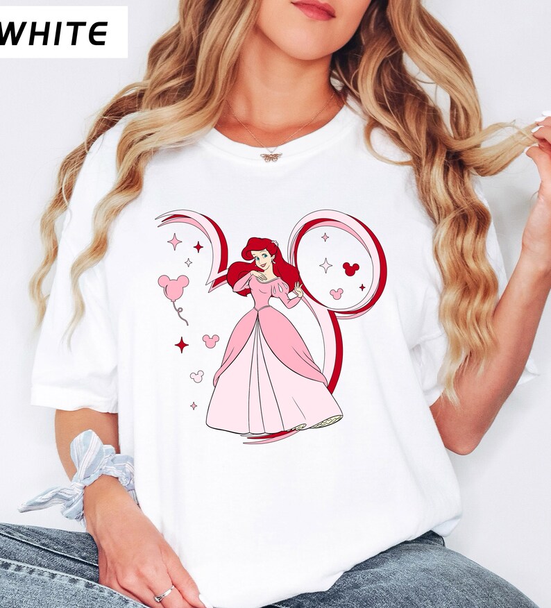 May include: White t-shirt featuring a cartoon illustration of a princess in a pink gown with red hair. The design includes a red outline of Mickey Mouse ears and pink heart-shaped balloons. The word "WHITE" is printed in the upper left corner.