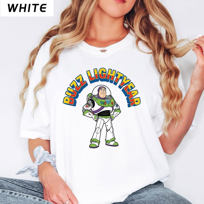 Op de afbeelding: Wit T-shirt met een afbeelding van Buzz Lightyear uit Toy Story. Het ontwerp bevat de woorden "BUZZ LIGHTYEAR" in een gebogen, veelkleurig lettertype boven het personage. Het personage is afgebeeld in zijn kenmerkende groene, witte en paarse ruimtepak.