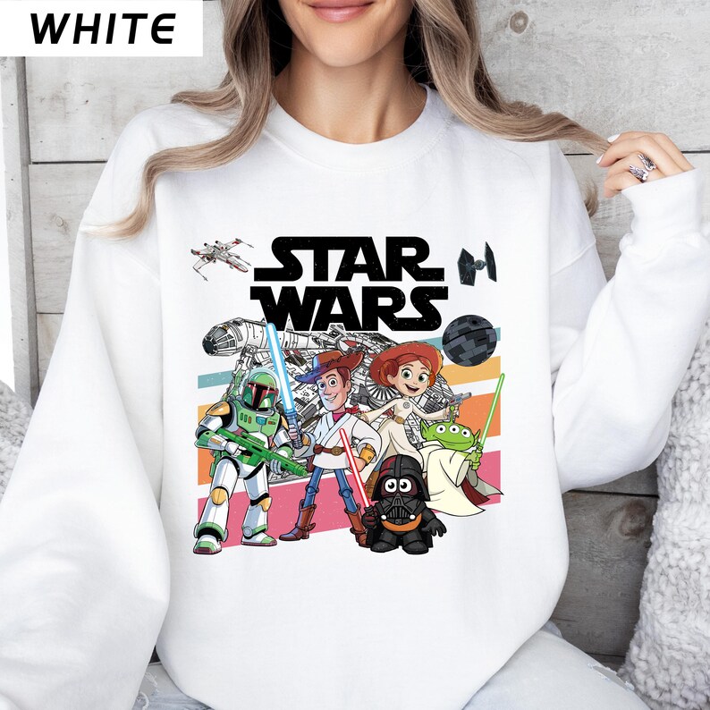 Puede incluir: Sudadera blanca con un dise&ntilde;o colorido de personajes de Star Wars y el texto "STAR WARS". El dise&ntilde;o incluye personajes como Boba Fett, Woody y Darth Vader, sobre un fondo a rayas.